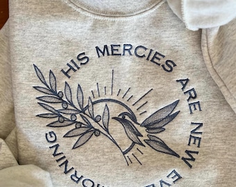 Op de afbeelding: Een grijs sweatshirt met een zwart geborduurd ontwerp. Het ontwerp toont een vogel, een tak met bladeren en een zon. De tekst rond het ontwerp luidt "Zijn genaden zijn elke morgen nieuw".