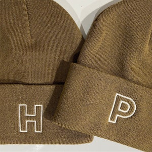 May include: Two brown knit beanies with white embroidered letters. The beanie on the left has the letter "H" embroidered on it. The beanie on the right has the letter "P" embroidered on it.