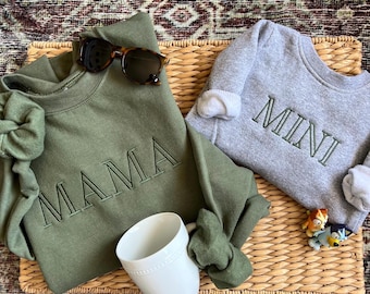 May include: Two green and gray sweatshirts with the words "MAMA" and "MINI" embroidered on them. The sweatshirts are laying on a woven basket with a white mug and two small toys.