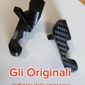 May include: Two black, 3D-printed automotive parts, one with a carbon fiber pattern and the other with an emblem. The text "Gli Originali" and "Diffidate dalle imitazioni" is displayed in red.