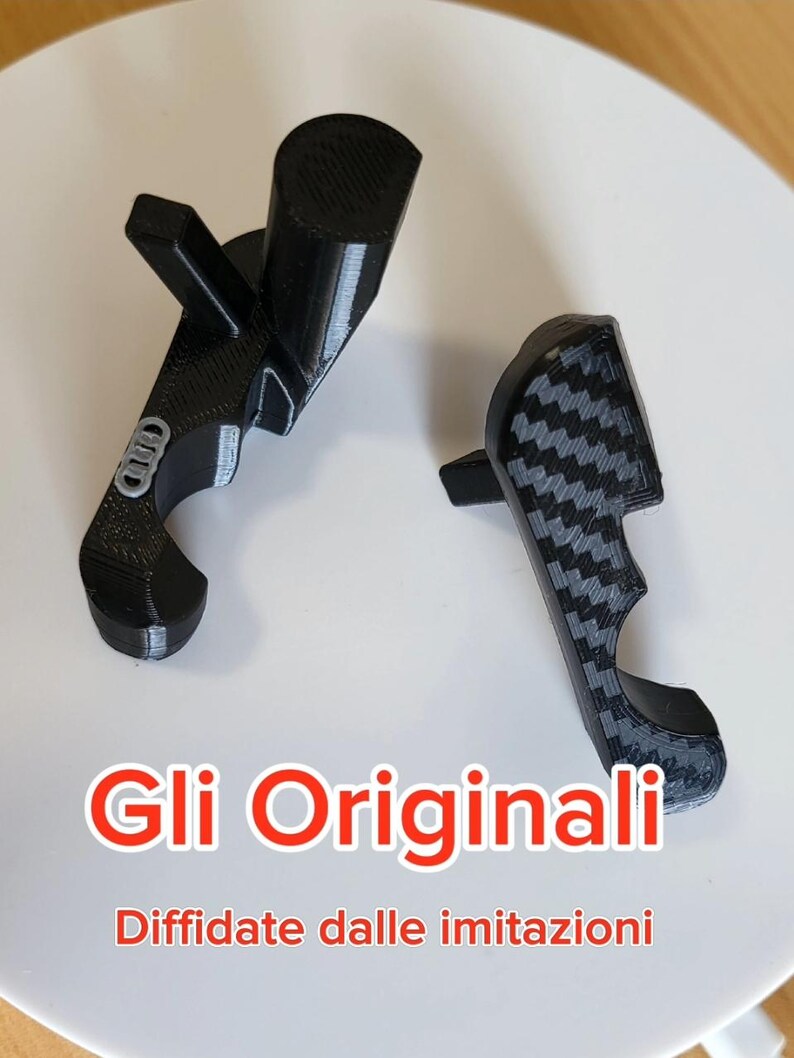May include: Two black, 3D-printed automotive parts, one with a carbon fiber pattern and the other with an emblem. The text "Gli Originali" and "Diffidate dalle imitazioni" is displayed in red.