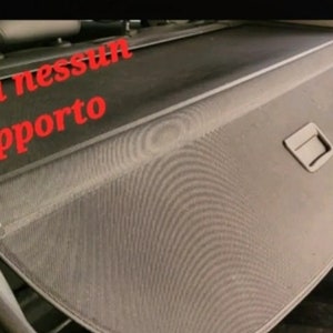 May include: A black car cargo cover with a mesh design. The cover has a rectangular shape with a handle and is installed in the back of a vehicle. Red text in Italian reads "senza nessun supporto".