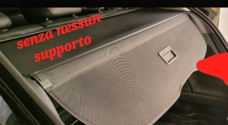 May include: A black car cargo cover with a mesh design. The cover has a rectangular shape with a handle and is installed in the back of a vehicle. Red text in Italian reads "senza nessun supporto".