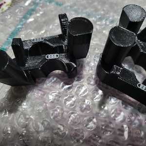 May include: Two black 3D-printed components with a cylindrical and rectangular design. Each component has a curved section with a small, white logo. The items are resting on bubble wrap.