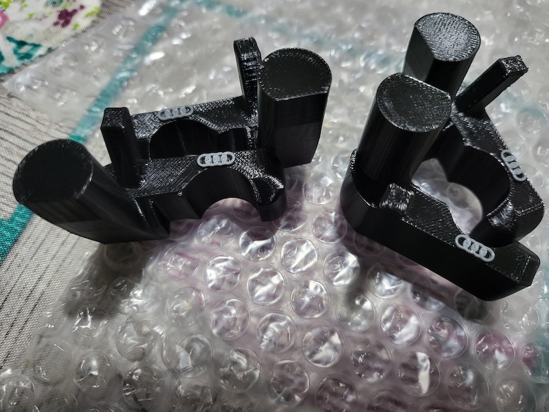 May include: Two black 3D-printed components with a cylindrical and rectangular design. Each component has a curved section with a small, white logo. The items are resting on bubble wrap.
