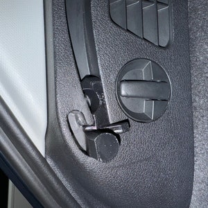 May include: Close-up of a black plastic car interior panel with a vent, a circular control, and a latch mechanism. The panel has a textured surface. The latch is open, revealing a metal component.