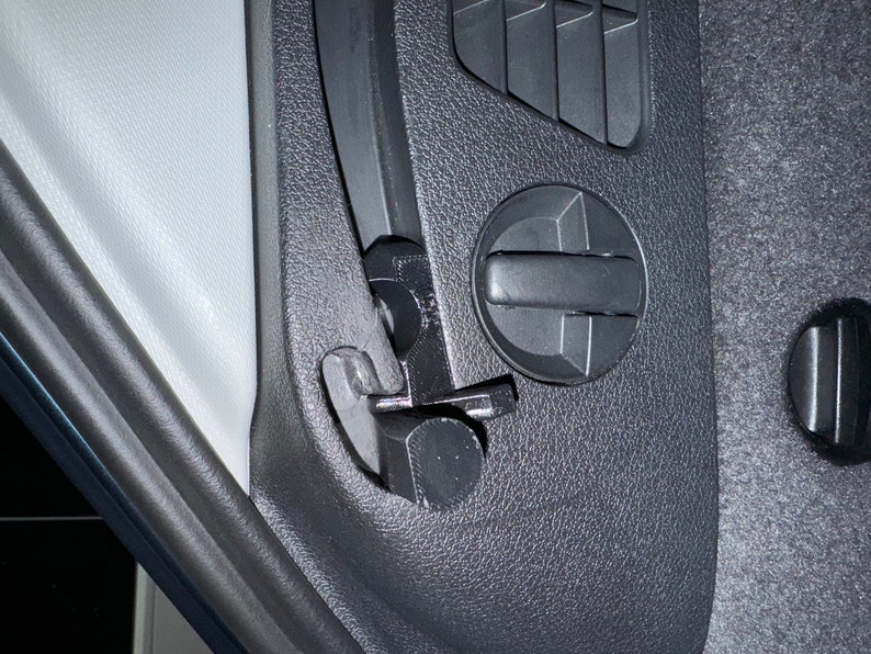 May include: Close-up of a black plastic car interior panel with a vent, a circular control, and a latch mechanism. The panel has a textured surface. The latch is open, revealing a metal component.