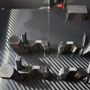 May include: Four black 3D-printed automotive parts with a white logo, arranged on a textured, dark gray surface. The parts have a distinctive shape with cylindrical and rectangular elements. A small, rectangular object is also visible.