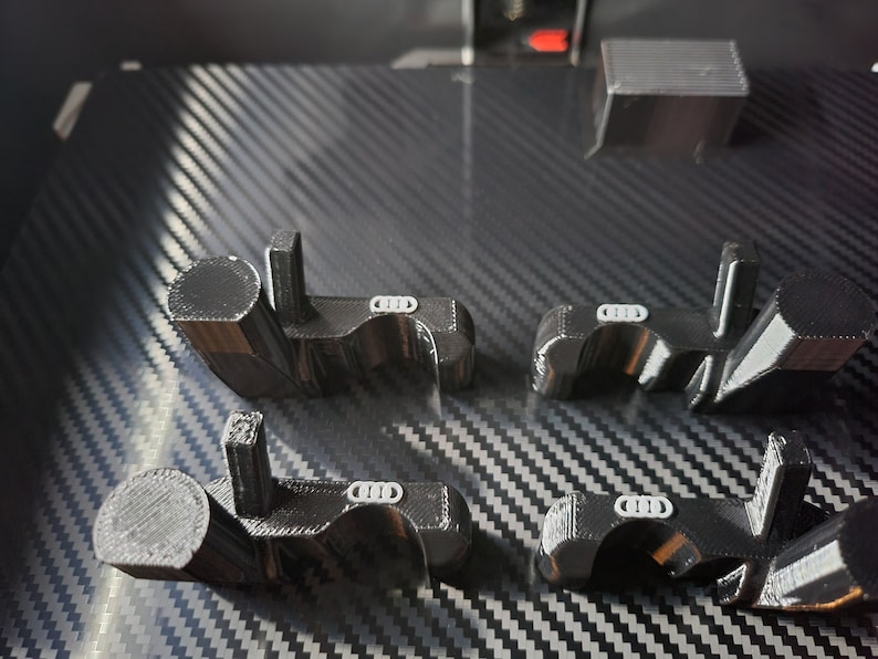 May include: Four black 3D-printed automotive parts with a white logo, arranged on a textured, dark gray surface. The parts have a distinctive shape with cylindrical and rectangular elements. A small, rectangular object is also visible.