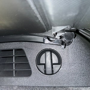 May include: Close-up of a black car interior featuring a vent, a circular handle, and a latch mechanism. The surfaces have a textured pattern. The image is taken from a low angle.