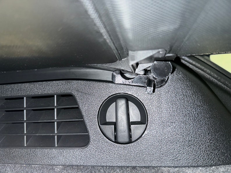 May include: Close-up of a black car interior featuring a vent, a circular handle, and a latch mechanism. The surfaces have a textured pattern. The image is taken from a low angle.