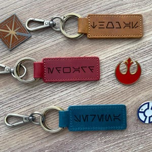 May include: Three keychains with leather straps and metal rings. The straps are engraved with different symbols. The first keychain has a brown strap with the symbol "HKOL", the second keychain has a red strap with the symbol "XOAF", and the third keychain has a blue strap with the symbol "KAI7VIM".