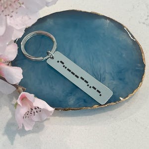May include: A silver-colored rectangular keychain with a Morse code pattern engraved on it. The keychain is attached to a silver ring. The background is a blue, agate-style coaster with a gold edge, and pink flowers.