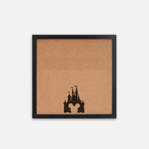 May include: Square corkboard with a black frame. A black silhouette of a castle with Mickey Mouse ears is centered at the bottom. The corkboard is a light brown color.