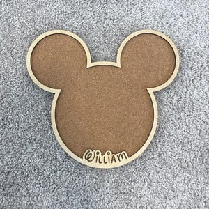 May include: A wooden cork board in the shape of Mickey Mouse's head. The board has a light brown cork center and a light wood border. The name "William" is carved in cursive at the bottom.