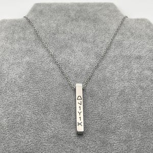 May include: A silver-colored necklace with a vertical rectangular pendant. The pendant has etched symbols and is suspended from a delicate chain. The necklace is displayed on a gray surface.