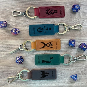 May include: Five leather keychains with personalized names, each with a unique design and a silver key ring. The keychains are displayed on a wooden surface with dice.