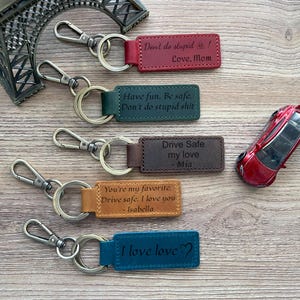 May include: Five leather keychains with different engraved messages. The keychains are brown, green, blue, and red. The messages include "Don't do stupid! Love. Mom", "Have fun. Be safe. Don't do stupid shit", "Drive Safe my love - Mia", "You're my favorite. Drive safe. I love you - Isabella", and "I love love".