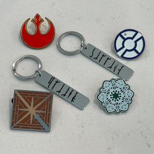 May include: Collection of pins and keychains with various designs. Includes a red and gold emblem pin, two keychains with etched text, a blue and white circular pin, a square pin with a geometric design, and a green and white floral pin.