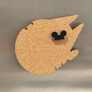 May include: A cork magnet shaped like the Millennium Falcon spaceship, with a black Mickey Mouse head pin attached. The magnet is light brown and has a textured surface. The pin is black with a silver outline.