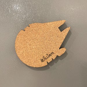 May include: A cork coaster shaped like the Millennium Falcon spaceship. The coaster is brown with black accents and has the name "William" written on it in black script.