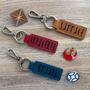 May include: A collection of keychains and pins. The keychains have leather tags in brown, red, and teal, each with black text. The pins include a geometric design, a red emblem, and a white and blue emblem. All items have metal hardware.