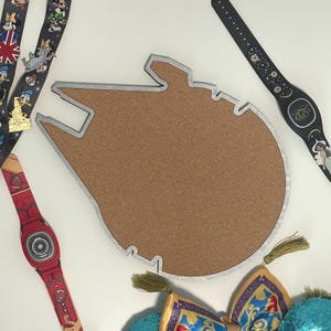 May include: A Millennium Falcon shaped cork display board with a silver-colored frame. The board is surrounded by various character-themed lanyards, a black wristband with moon and star patterns, a red wristband, and a pair of sequined ears.