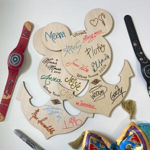 May include: Two light-colored wooden cutouts shaped like Mickey Mouse heads with anchor bases. The wood is covered in various colorful character signatures and names including Moana, Mickey Mouse, Pluto, Elsa, Snow White, Anna, Jessie, Chip, Woody, Goofy, Mr. Incredible, Mrs. Incredible, Donald Duck, and Lilo.