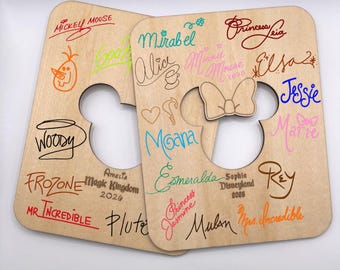 May include: Two wooden Mickey Mouse-shaped signs with character signatures in various colors. Signatures include Mickey Mouse, Woody, Frozone, Mirabel, Moana, and others. The signs have cut-out areas and are made of light-colored wood.