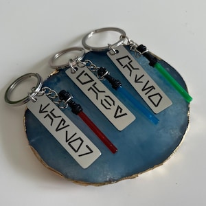 May include: Four keychains with lightsaber designs. Each keychain has a silver rectangular tag with a word in a stylized font. The lightsabers are red, blue, and green. The keychains are on a blue and gold geode.