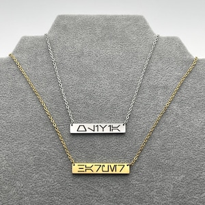 May include: Two necklaces with rectangular pendants. One is silver with the text "ΔN1Y1K", and the other is gold with the text "EK7UM7". Both necklaces are displayed on a gray surface.