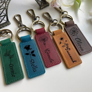 May include: A collection of personalized leather keychains in various colors, including green, blue, burgundy, tan, and gray. Each keychain features a floral design and a name, such as "Annie," "Stella," "Camila," "Savannah," and "Olivia."