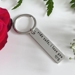 May include: Silver rectangular keychain with the text "ride safe, i love you" and a heart symbol. The keychain is attached to a round key ring. The background includes a red rose and greenery.