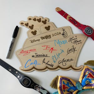 May include: A light-colored wooden cutout shaped like a cruise ship, featuring the text Disney Destiny 2026, Mickey Mouse ears, and various character signatures in black, red, and blue ink. A black marker, two wristbands, and a bow are nearby.