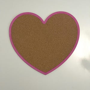 May include: A heart-shaped cork board featuring a smooth, light brown textured surface framed by a solid, vibrant pink border. The object is shaped like a classic heart with a pointed bottom and a curved top, providing a flat display area.