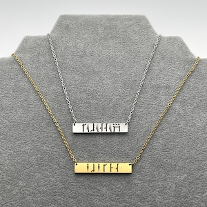May include: Two necklaces with bar pendants. One is gold-toned, the other silver-toned. Each pendant has a word cut out in a stylized font. The necklaces are displayed on a gray surface.