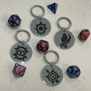 May include: A collection of silver keychains with engraved black designs, including a sunburst, a hand, and a compass. The keychains are accompanied by several polyhedral dice in shades of blue, red, and gold, perfect for tabletop gaming.