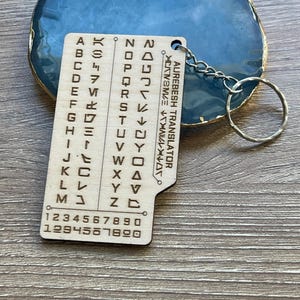 May include: A wooden Aurebesh Translator keychain with a silver keyring. The keychain features engraved Aurebesh characters, numbers, and the text "AUREBESH TRANSLATOR". The keychain is light brown and rests on a blue and gold surface.