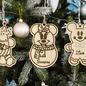 May include: Three wooden Christmas ornaments shaped like cartoon characters, hanging on a Christmas tree. The ornaments are light brown with dark brown outlines and details. Each ornament has a name: Sophia, Serena, and Liam.