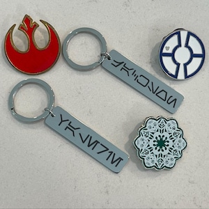 May include: A collection of Star Wars themed accessories. Includes a red and gold emblem pin, two keychains with gray tags, and two round pins. The keychains have gray rings and text on the tags. One pin is white and blue, and the other is green and white.