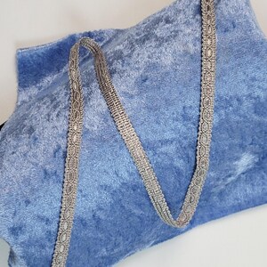 May include: A blue velvet bag with a silver-colored decorative trim. The bag has a soft, plush texture and a simple design. The trim is a delicate, patterned ribbon that serves as a strap.