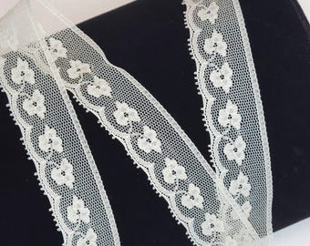 May include: White lace trim with a delicate floral pattern. The trim is rolled up on a black surface.