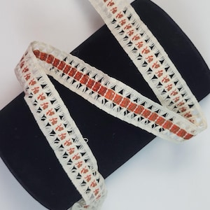 Op de afbeelding: Crèmekleurig kanten lint met een herhalend bloemmotief in roestoranje. Het lint is gedrapeerd over een zwart, cilindrisch object, dat het ingewikkelde opengewerkte ontwerp laat zien. De randen van het lint hebben een geschulpte rand.