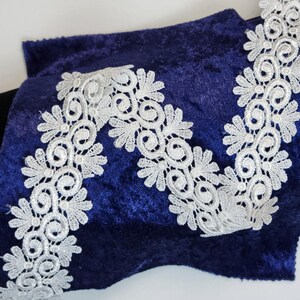 Puede incluir: Primer plano de tela de terciopelo azul marino con ribete de encaje blanco. El encaje presenta un diseño floral con espirales y formas de pétalos. La tela está doblada, revelando una tela negra debajo.