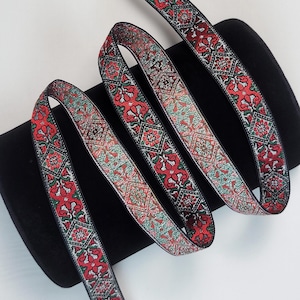 Puede incluir: Cinta decorativa con un patrón floral repetido en rojo, verde y negro. La cinta se muestra en una disposición en bucle sobre un fondo negro, mostrando su intrincado diseño y paleta de colores. Ideal para manualidades o decoración.