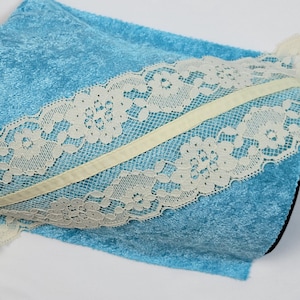 Pode incluir: Um tecido de veludo azul claro com uma borda de renda branca. A renda tem um padrão floral e é acentuada por uma fita branca fina.