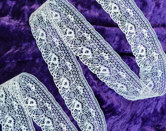 May include: A length of white lace ribbon with a detailed floral design, set against a rich purple velvet backdrop. The ribbon features an intricate openwork pattern, suitable for crafts and decorative purposes. This ribbon is perfect for adding a touch of sophistication to any project.