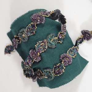 Puede incluir: Una diadema con cuentas con un diseño floral. La diadema presenta cuentas y lentejuelas moradas, verde azulado y doradas. La diadema se muestra sobre un trozo de tela verde.