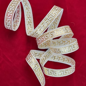 Puede incluir: Cinta blanca con un diseño de greca dorada. La cinta está enrollada sobre un fondo rojo. El patrón dorado tiene un brillo metálico. Adecuada para manualidades y envolver regalos.