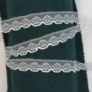Puede incluir: Borde de encaje blanco con borde festoneado y patrón floral. El encaje se muestra sobre un fondo de tela verde oscuro.